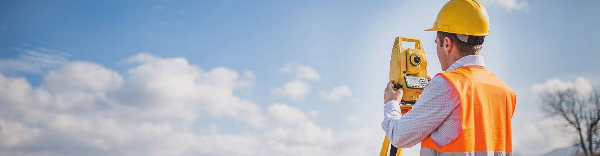Homem usando colete de seguran&ccedil;a laranja em uma estrada, analisando o terreno atrav&eacute;s do equipamento.